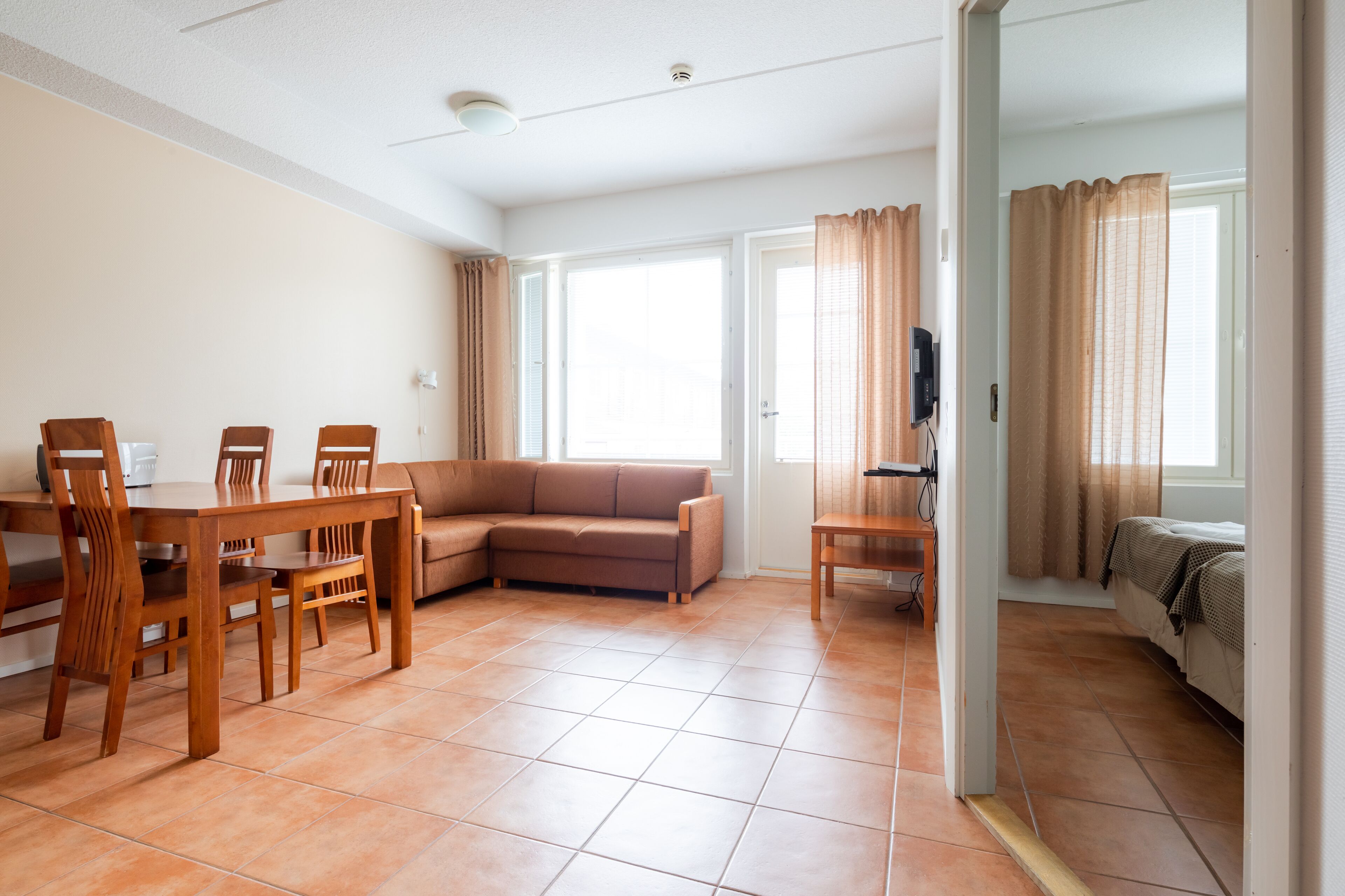 A spacious living area with a dining table, sofa, and chairs. There are large windows with curtains, a television, and a door leading to another room.