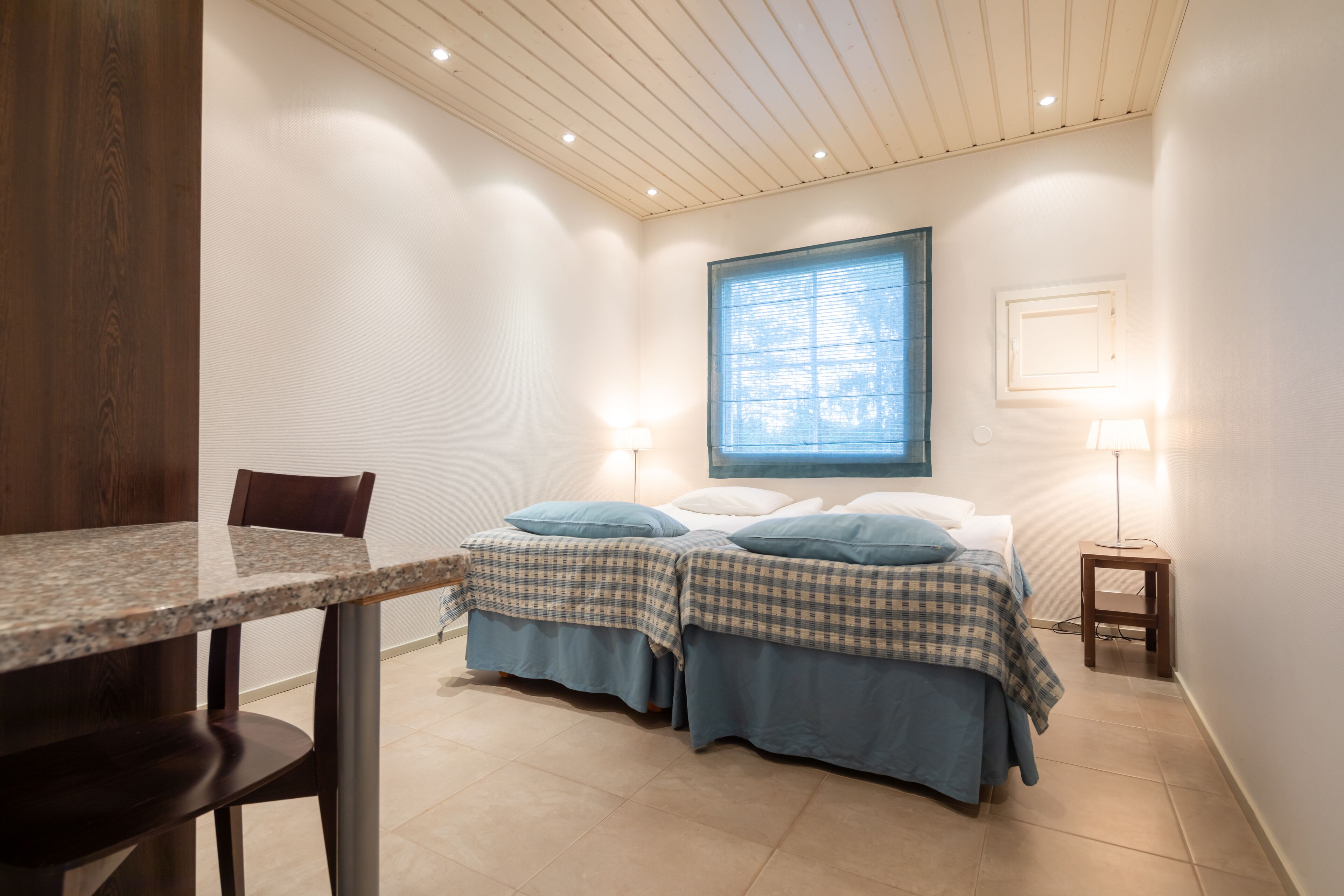 A bedroom with two beds, a dining table, and a window with blinds.
