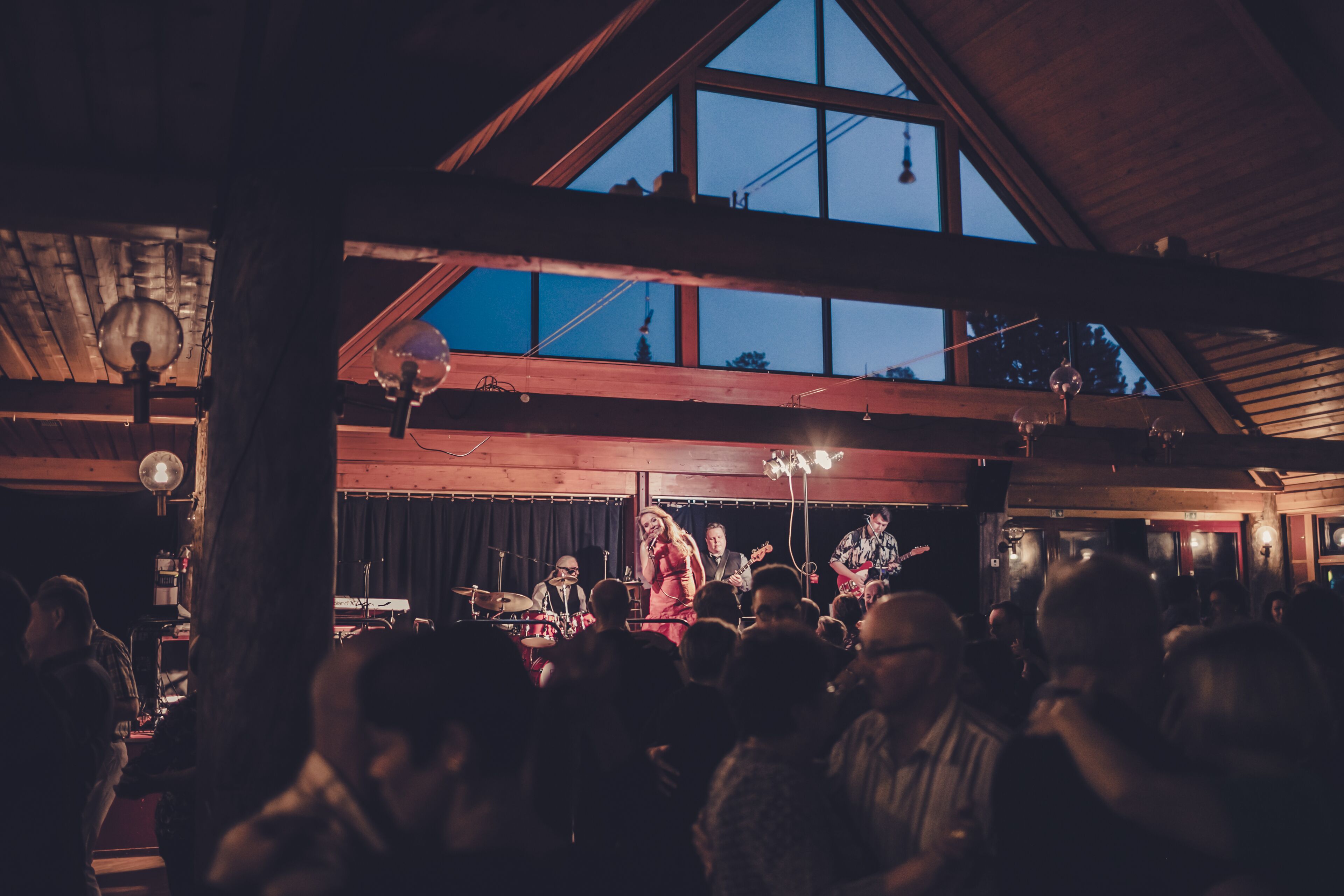 Un groupe de musique se produit sur une scène devant un public, dans un lieu doté de poutres en bois et de grandes fenêtres.