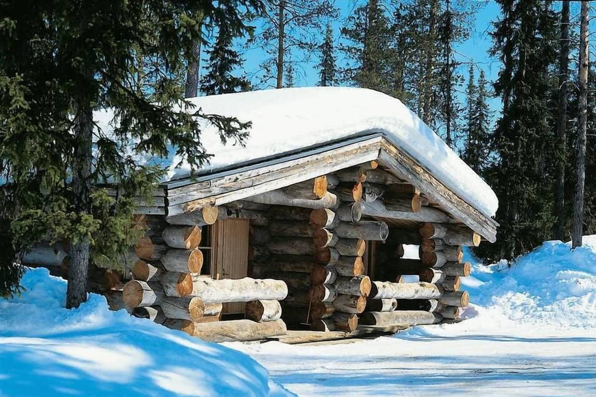 Una cabaña de troncos con techo nevado, rodeada de nieve y árboles.