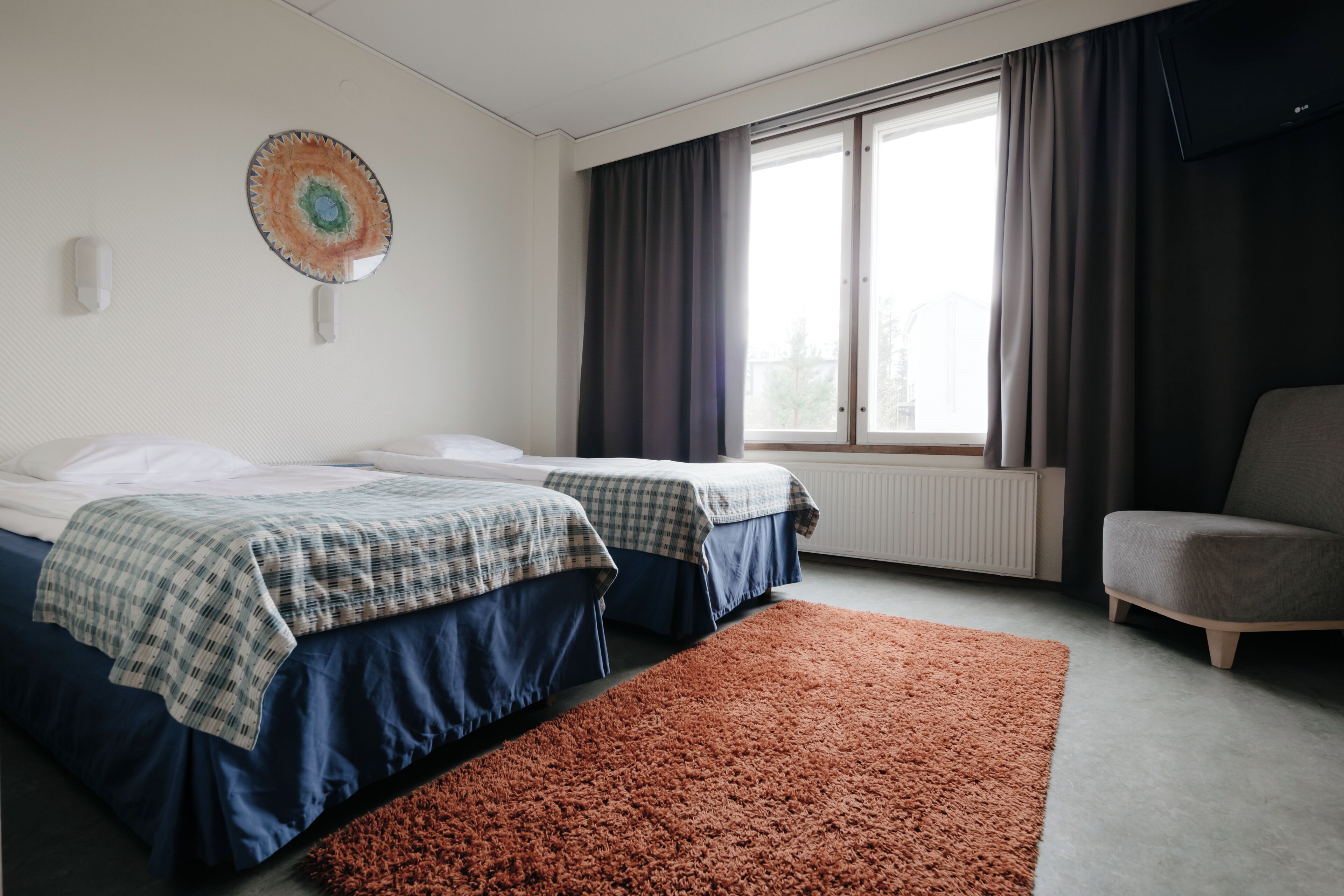 Habitació d'hotel amb dos llits, una finestra gran amb cortines, un radiador, una cadira i una decoració mural.