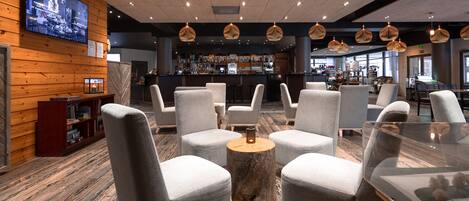A modern lounge area with grey upholstered chairs, a central wooden table, and a bar in the background.