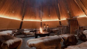 A cozy interior with a central fire, wooden walls, and fur-covered benches. - Lapland Hotels Ylläskaltio (Kolari)