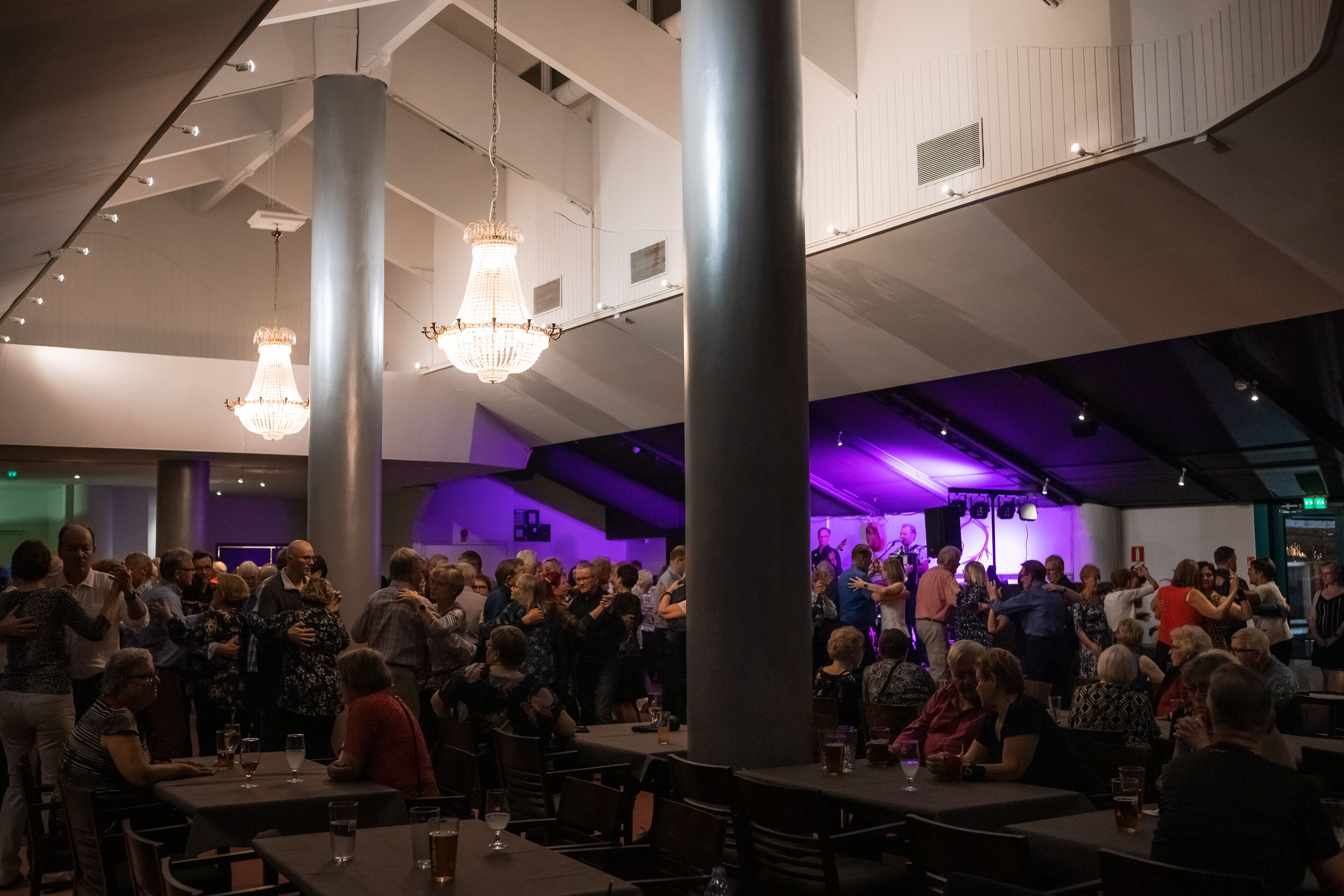 A crowded indoor venue with tables and chairs, large columns, and hanging chandeliers.