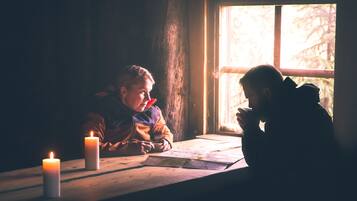 Dos niños sentados a una mesa de madera junto a una ventana con velas.