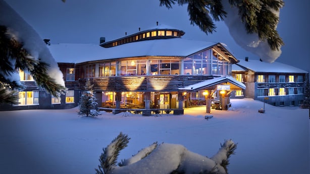 A large, multi-story lodge with a snow-covered roof and illuminated windows, surrounded by snow and pine trees. - Lapland Hotels Ylläskaltio (Kolari)