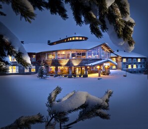 A large, multi-story lodge with a snow-covered roof and illuminated windows, surrounded by snow and pine trees.