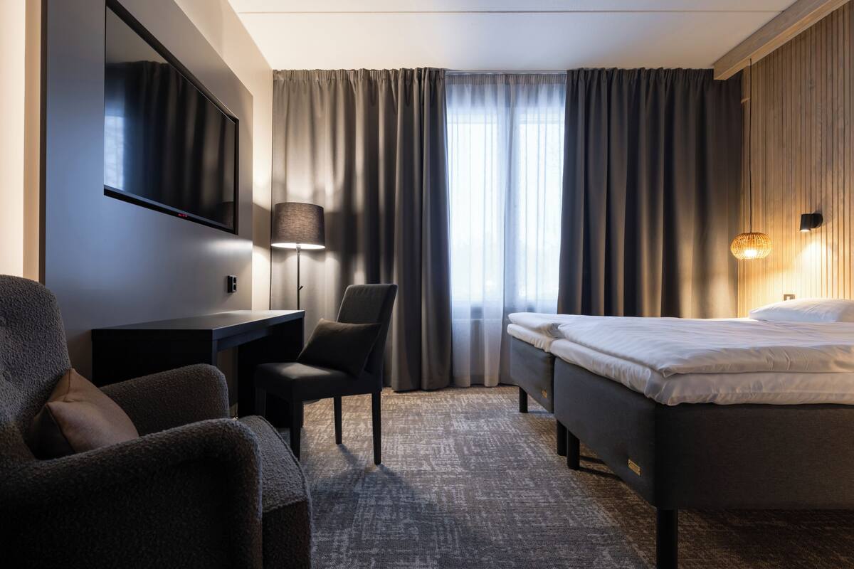 Habitación de hotel con cama, escritorio, silla, televisor y ventana con cortinas.