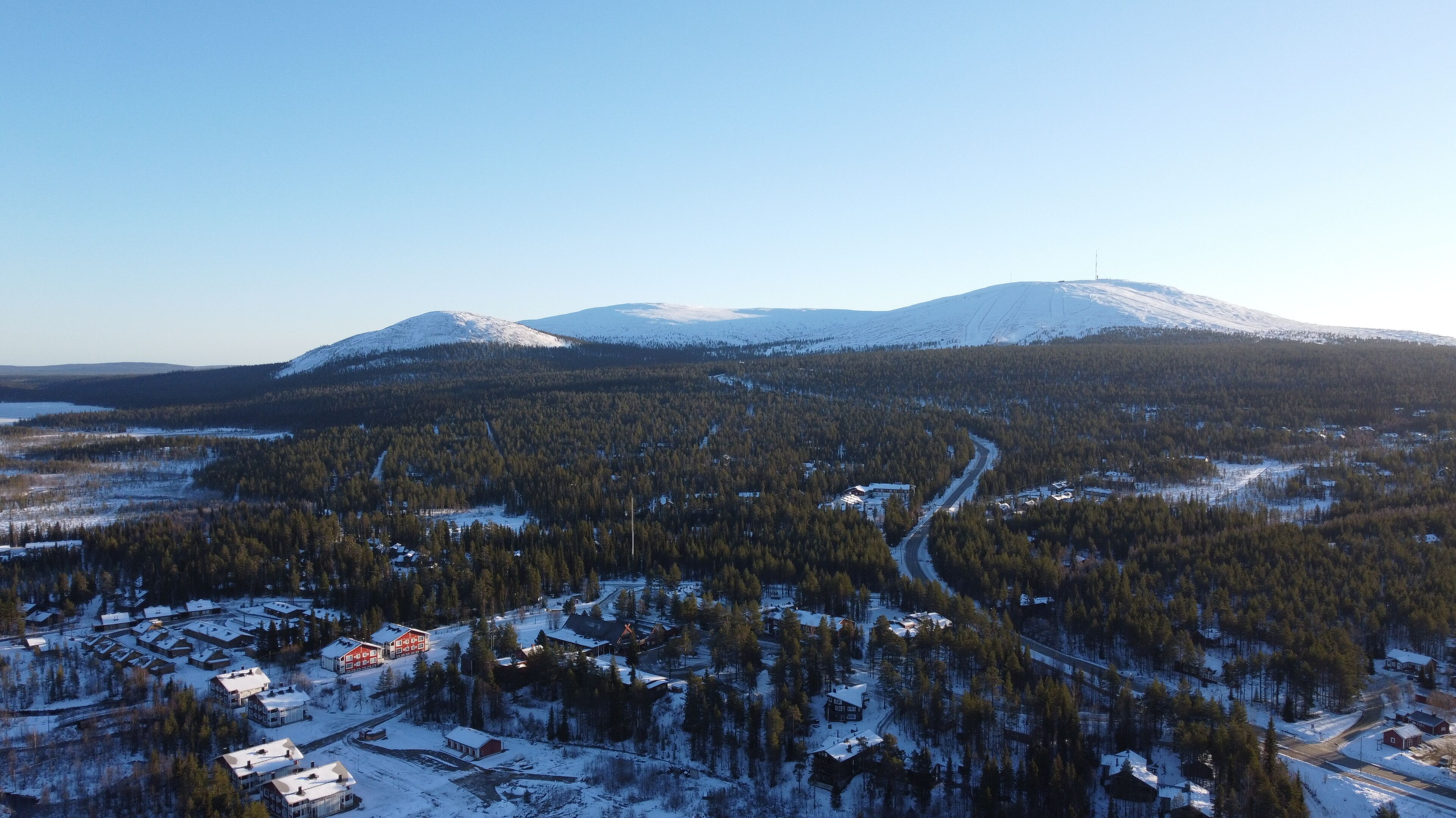 Ett flygbildsbild av ett snötäckt landskap med ett berg, en tät skog och en liten bebyggelse med några byggnader.