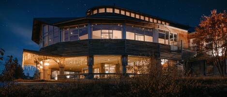 A modern, multi-level house with large windows and a wrap-around porch, surrounded by natural landscape.