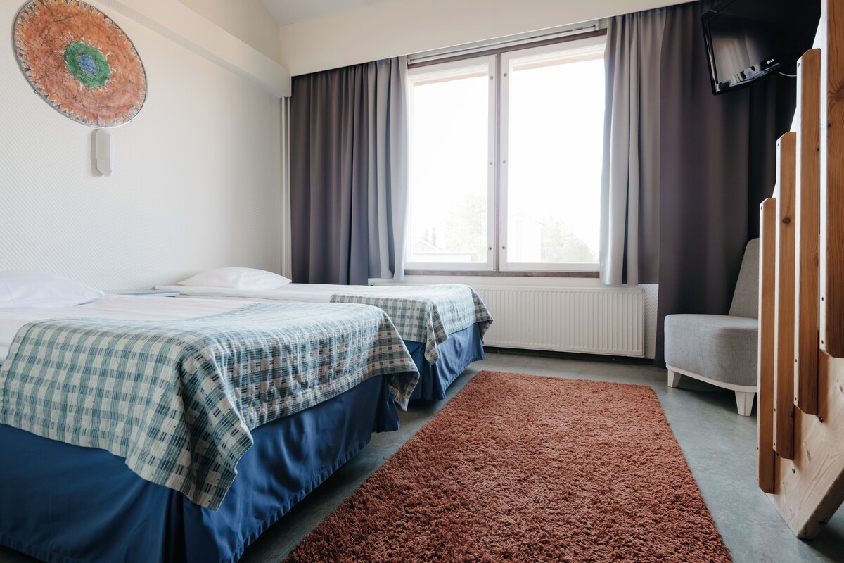 A hotel room with two beds, a large window with curtains, a TV, and a wooden chair.