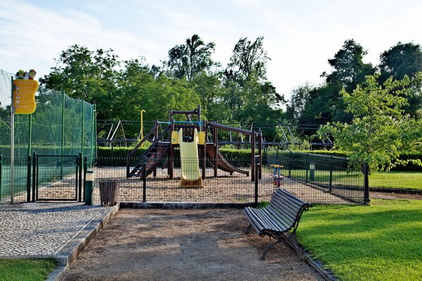 Un parque infantil con tobogán y estructuras para escalar, rodeado por una valla y bancos.