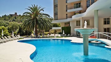 Piscina com espreguiçadeiras e uma fonte, rodeada por um edifício com varandas e vista para um castelo ao fundo.