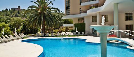 A swimming pool with lounge chairs and a fountain, surrounded by a building with balconies and a view of a castle in the background.