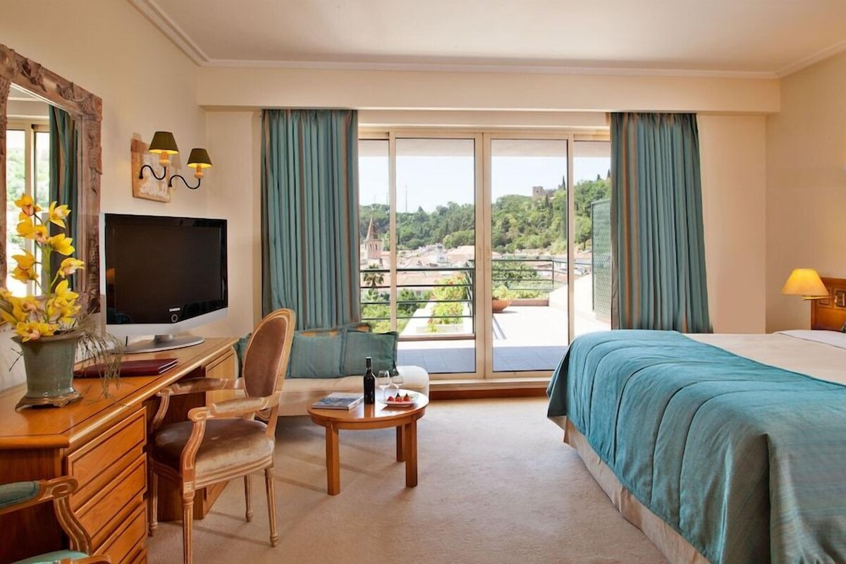 A hotel room with a bed, a desk with a chair, a television, and a balcony with curtains.