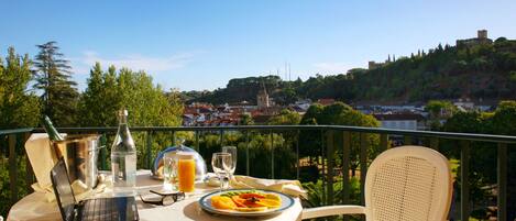 Uma mesa com um laptop, uma garrafa, um copo de sumo de laranja e uma travessa de fruta numa varanda com vista para uma cidade, uma colina e um castelo.