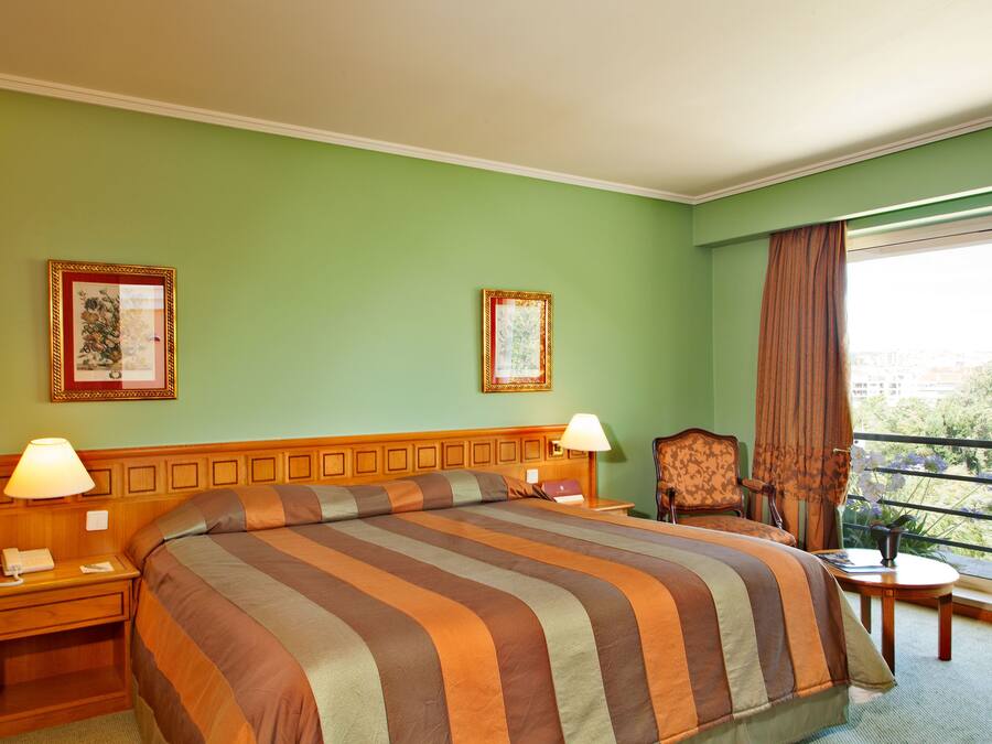 A hotel room with a large bed, two bedside tables with lamps, a chair, a small table, and two framed pictures on the wall.