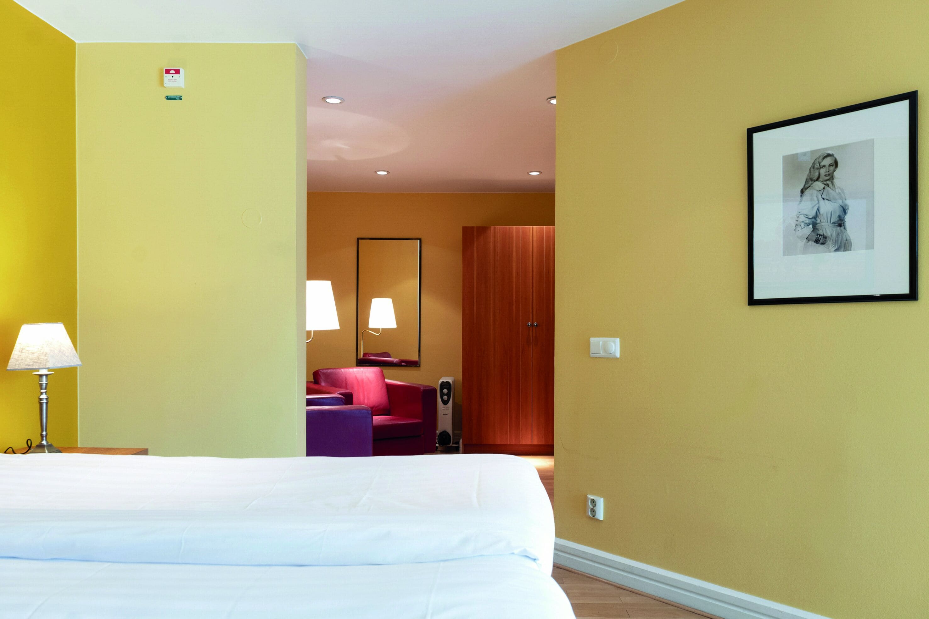 A hotel room with a large bed, a desk with a lamp, a red chair, a wooden wardrobe, and a framed picture on the wall.