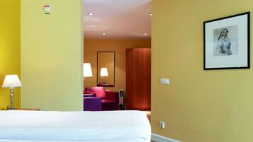 A hotel room with a large bed, a desk with a lamp, a red chair, a wooden wardrobe, and a framed picture on the wall.