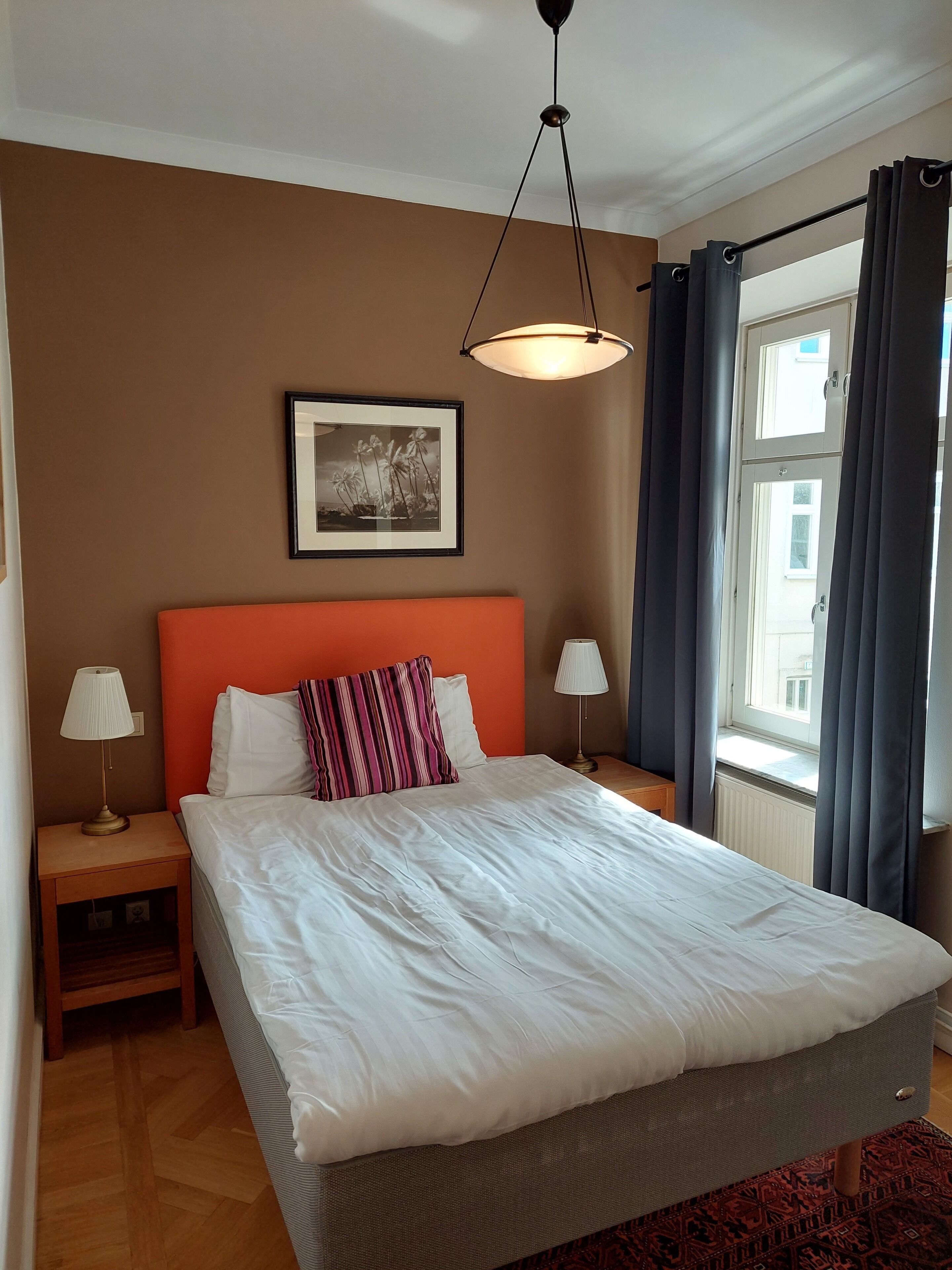 Un dormitorio ordenado con una cama grande, una mesita de noche con lámpara, un cuadro enmarcado en la pared y una ventana con cortinas.