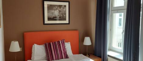 Un dormitorio ordenado con una cama grande, una mesita de noche con lámpara, un cuadro enmarcado en la pared y una ventana con cortinas.