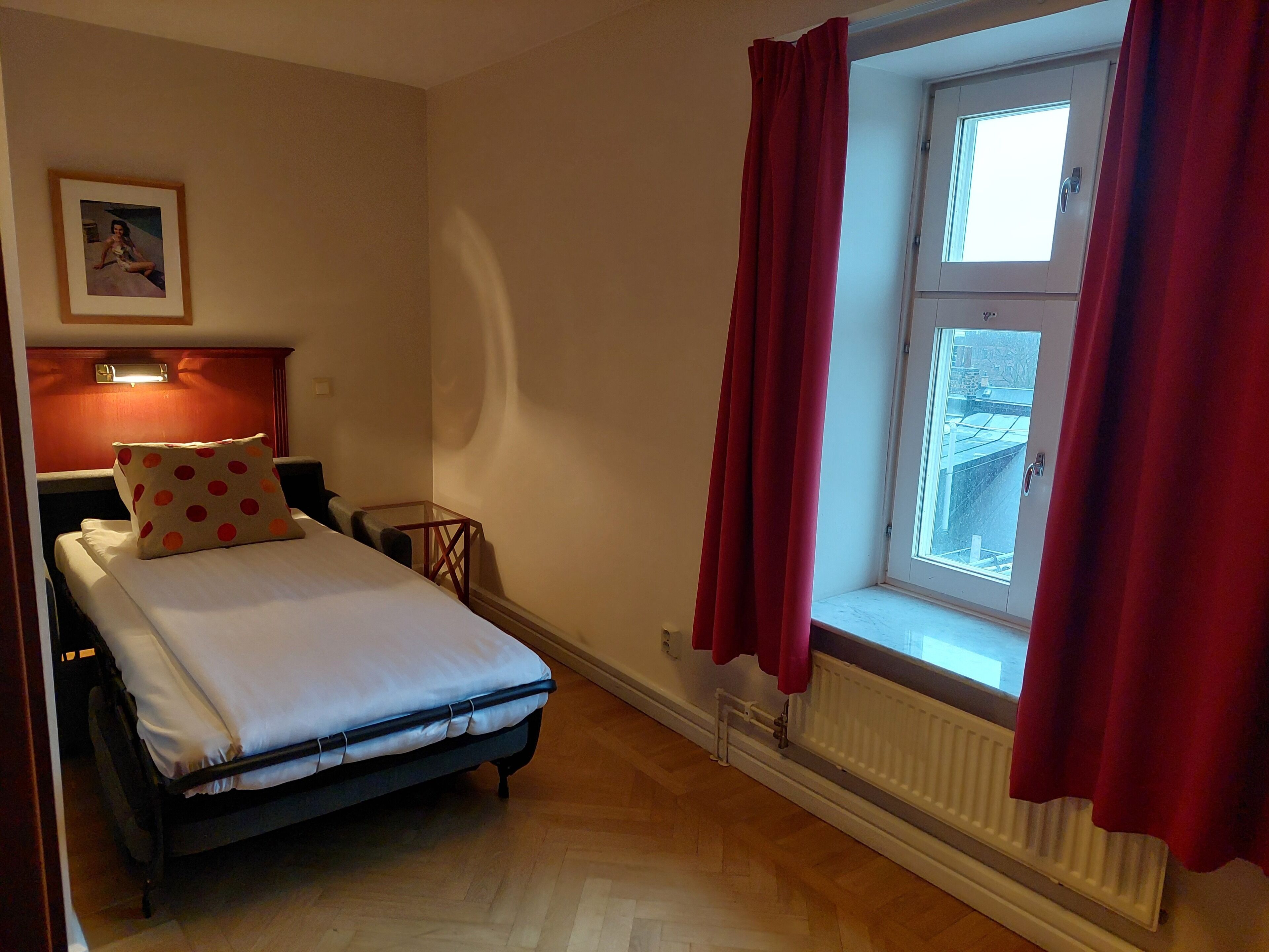 A bedroom with a bed, a nightstand, a red curtain, a window, and a radiator.