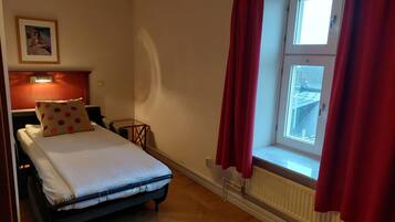 A bedroom with a bed, a nightstand, a red curtain, a window, and a radiator.