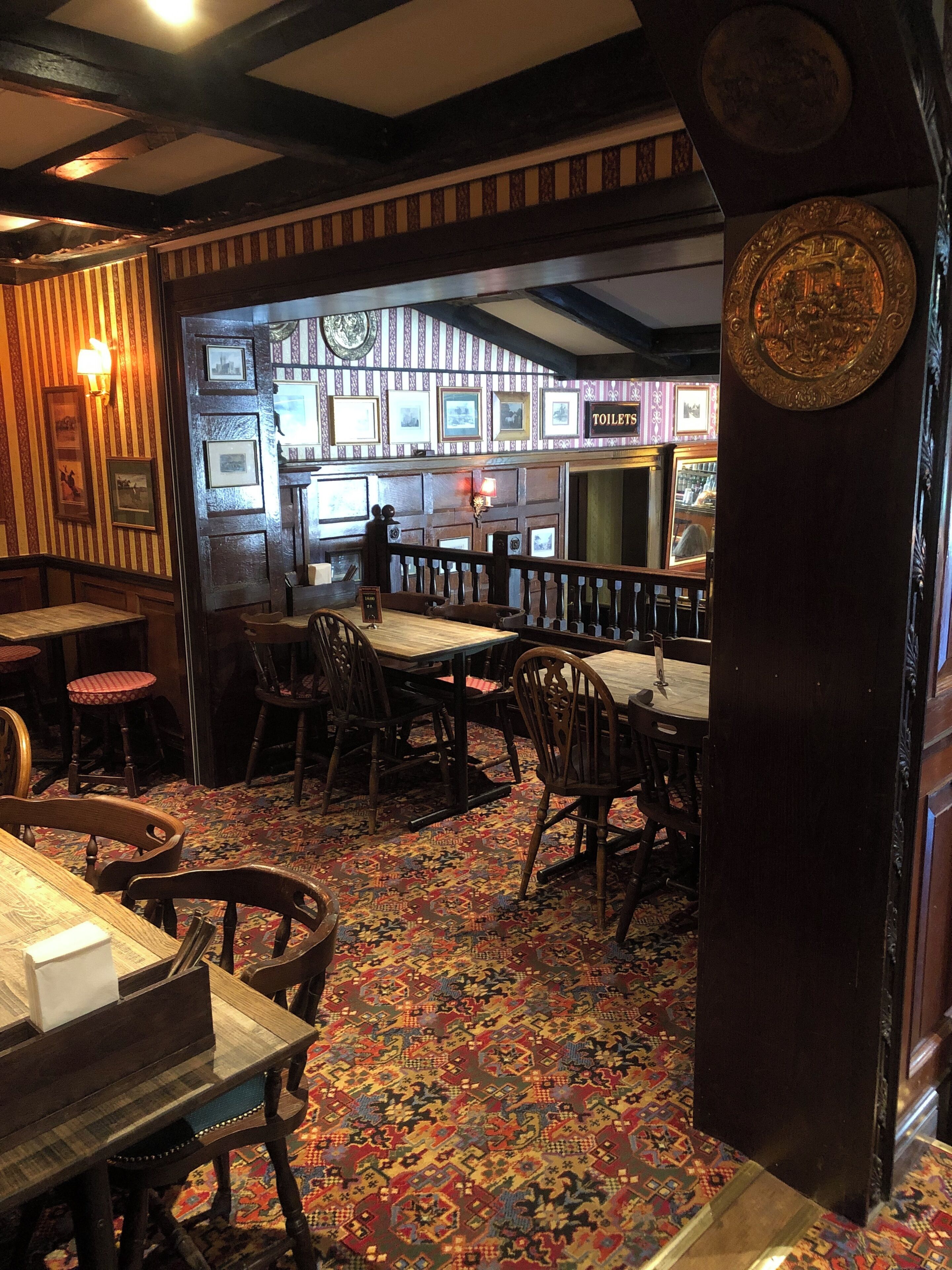 A traditional pub interior with wooden furniture, patterned carpet, and framed pictures on the walls.