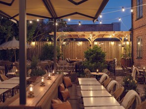 An outdoor patio with wooden tables and chairs, string lights, and a brick building in the background. - Hotel Bishops Arms Lund (Lund)