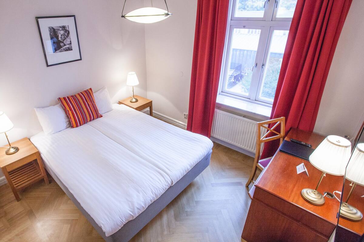 Un dormitorio ordenado con cama, lámparas de noche, una mesita de madera, un escritorio con lámpara, una silla y una ventana con cortinas rojas.