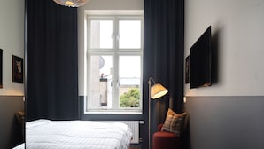 A bedroom with a large bed, a window with curtains, a floor lamp, a red ottoman, and a round table. - Hotel Bishops Arms Lund (Lund)