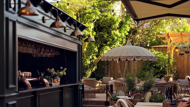 An outdoor seating area with wicker furniture, a bar with copper fixtures, and a large umbrella providing shade. - Hotel Bishops Arms Lund (Lund)