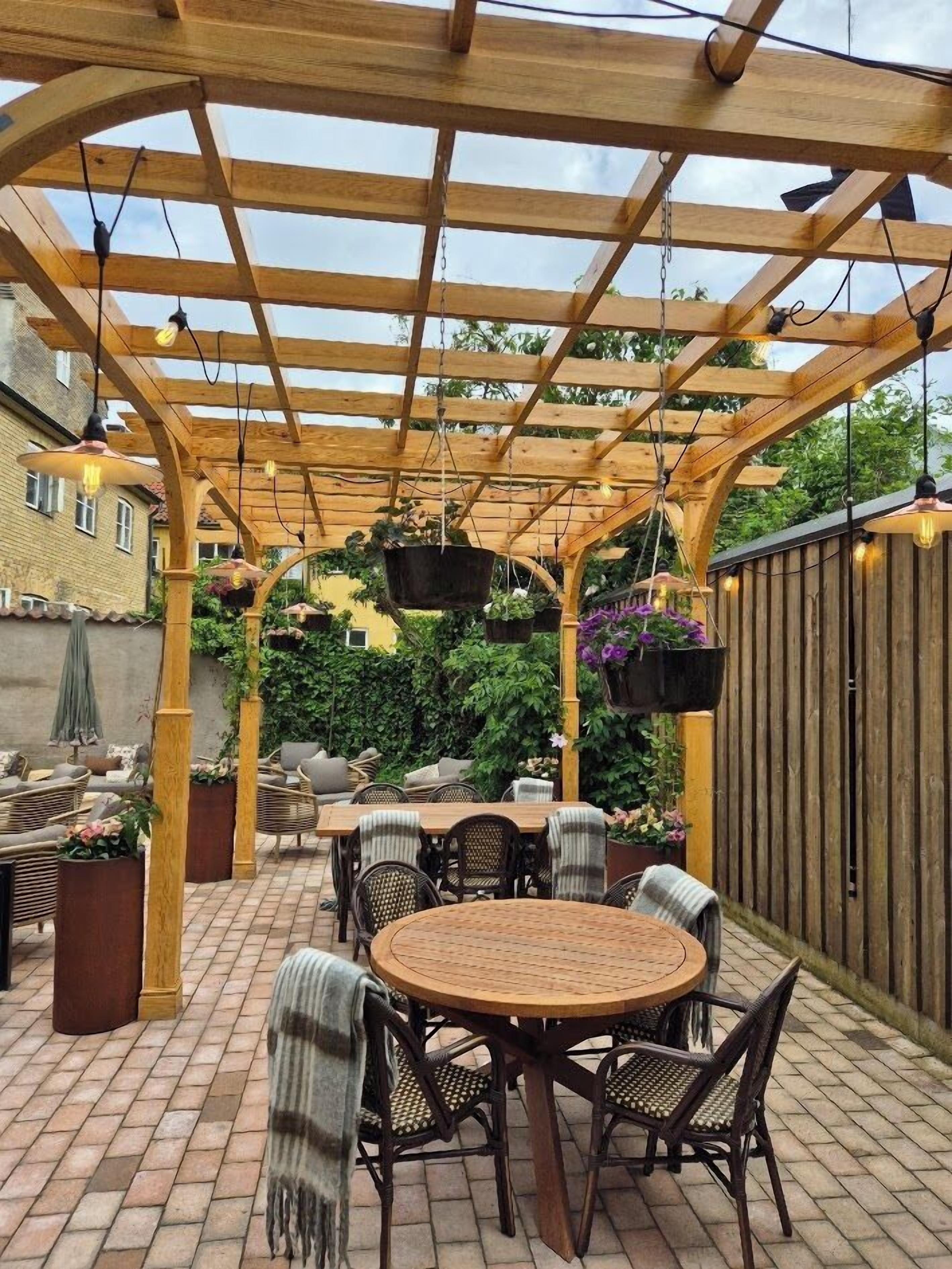 Een terras buiten met een houten pergola, ronde tafels, rieten stoelen, hangende plantenbakken en een tegelvloer.