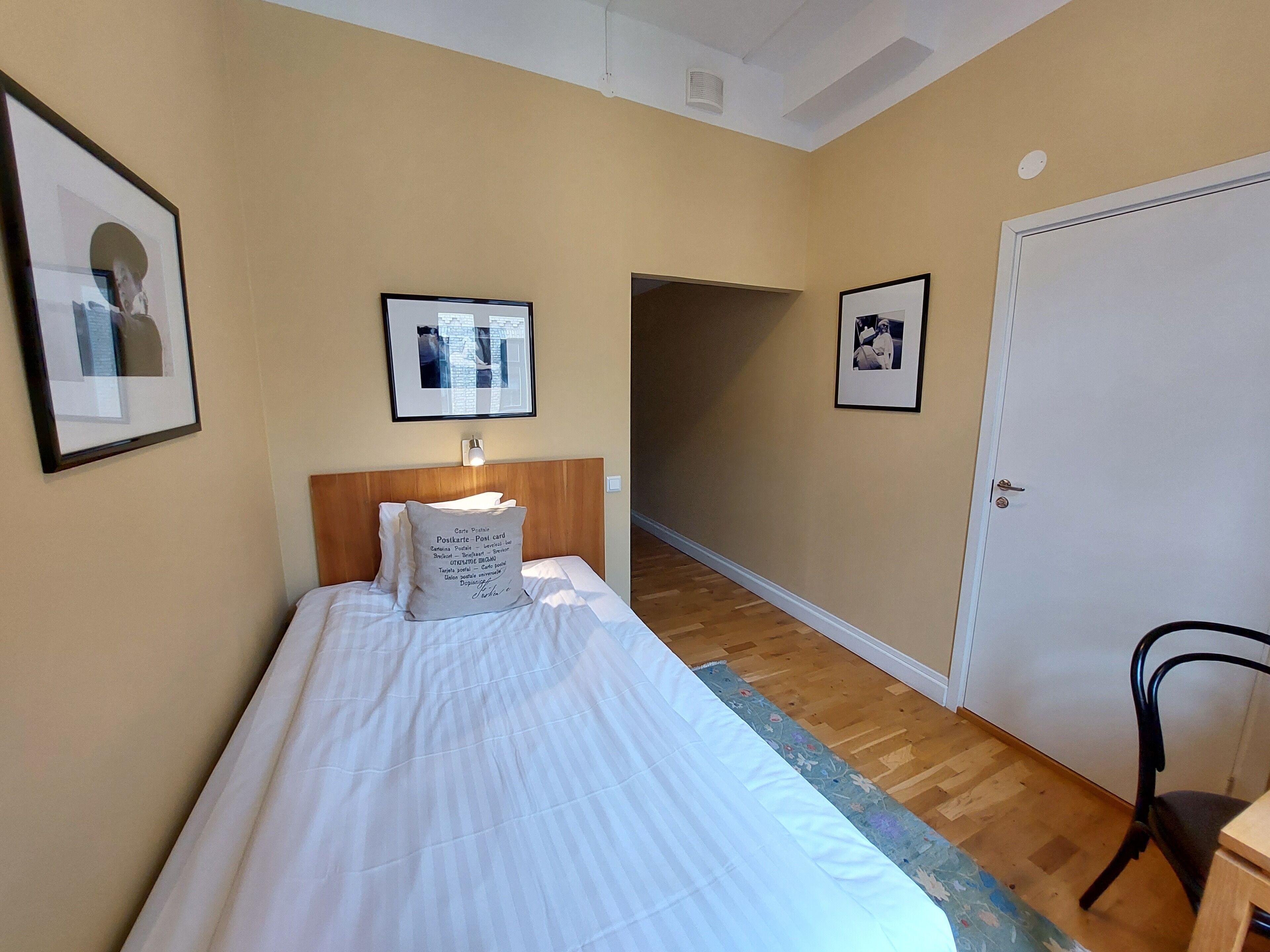 Een hotelkamer met een bed, een bureau en een stoel. Er hangen prenten in de wanden en een deur bevindt zich aan de rechterkant.