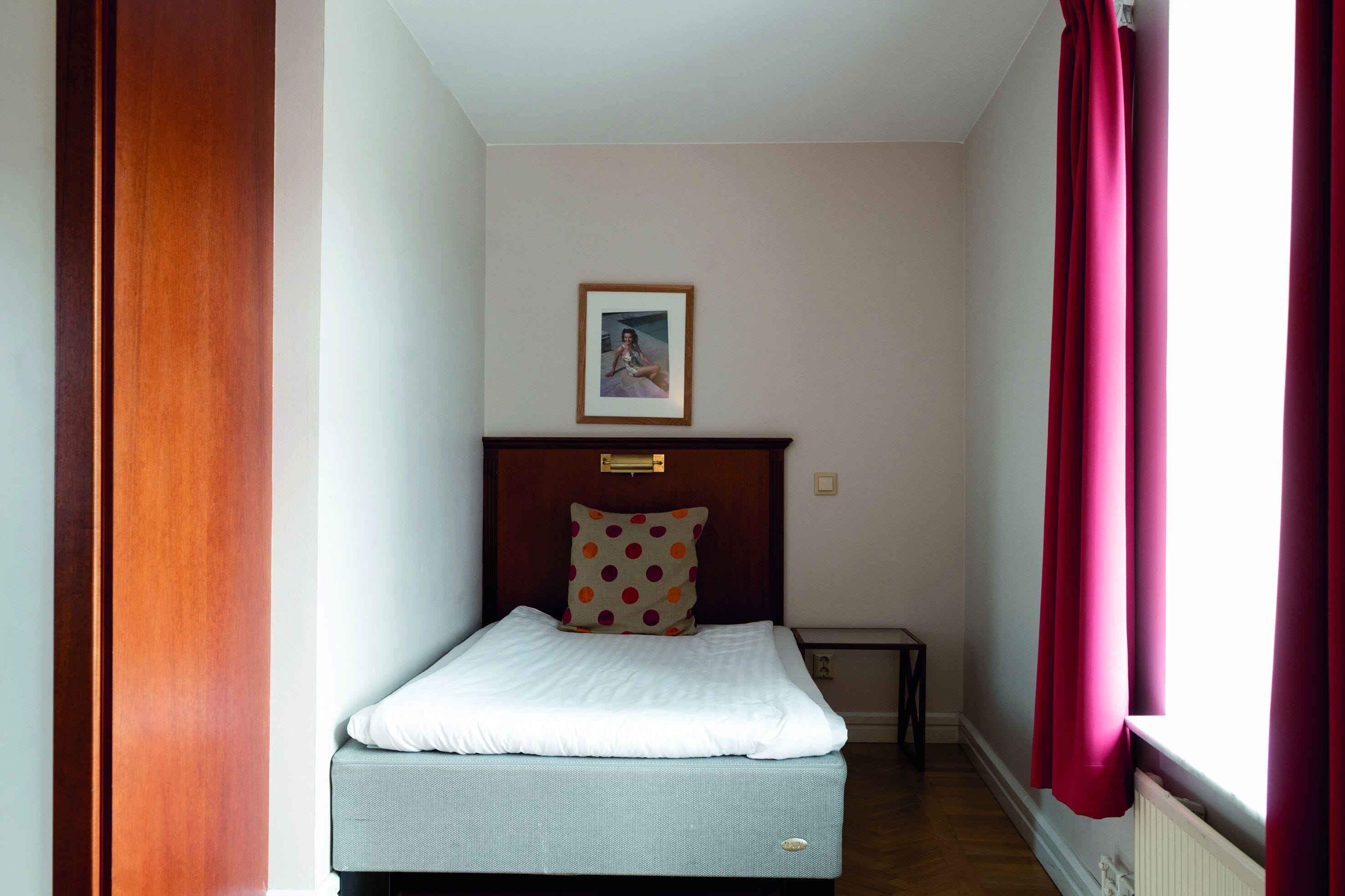 Een kleine, verzorgde slaapkamer met een bed, een houten hoofdbord, een ingelijst beeld aan de muur en een raam met rode gordijnen.