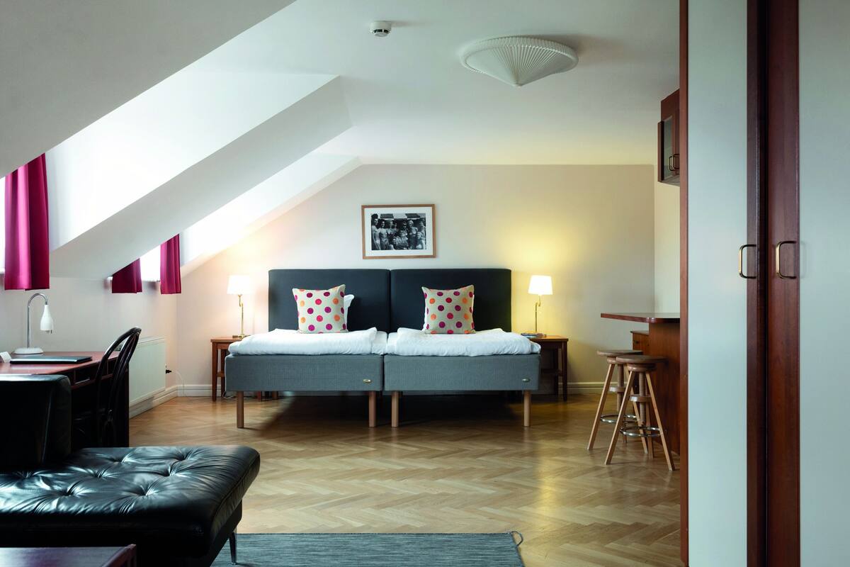 Un dormitorio moderno con techo estilo loft, una cama con dos almohadas, un escritorio con silla y un taburete.