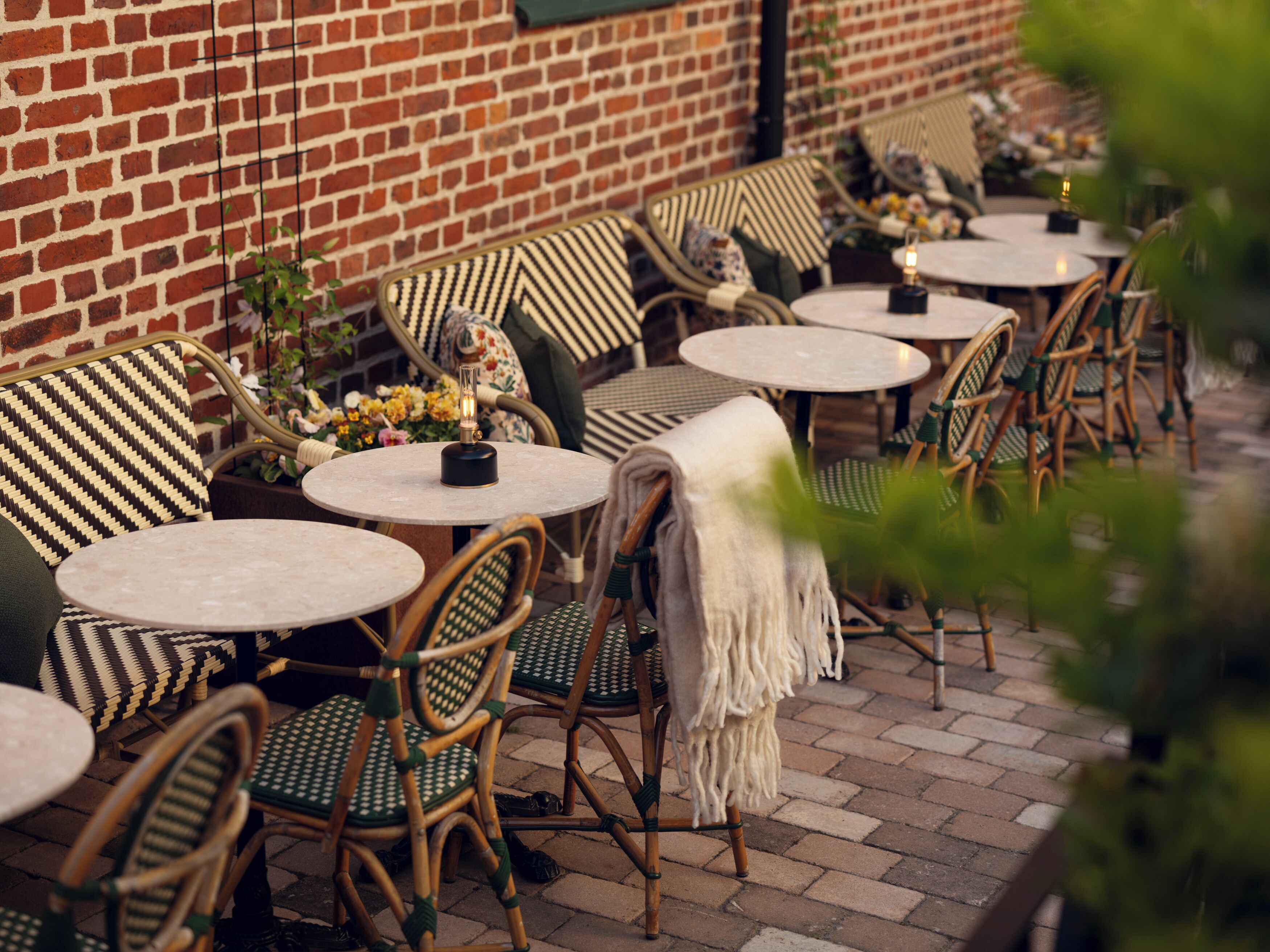 Outdoor seating with wicker chairs and round tables, a brick wall, and potted plants.