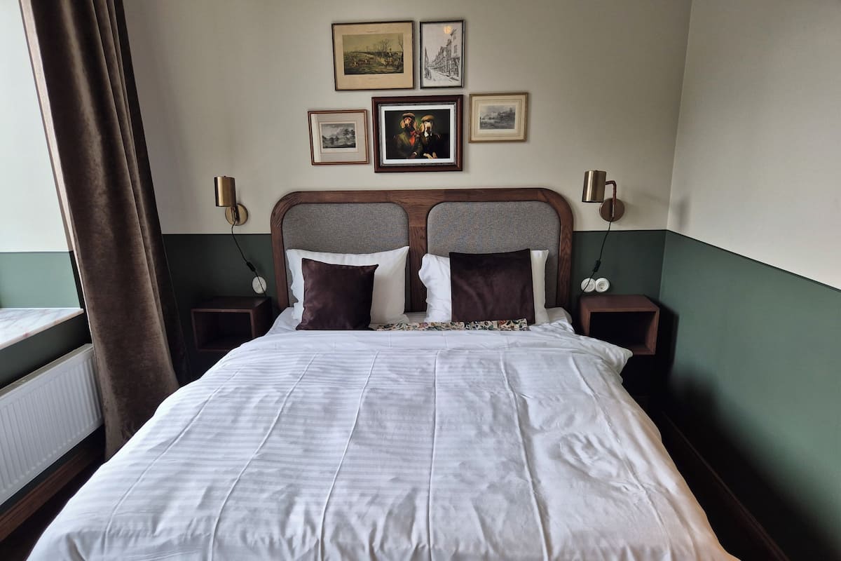 Una cama bien hecha con sábanas blancas y dos almohadas oscuras, un cabecero de madera y una mesita de noche con una lámpara.