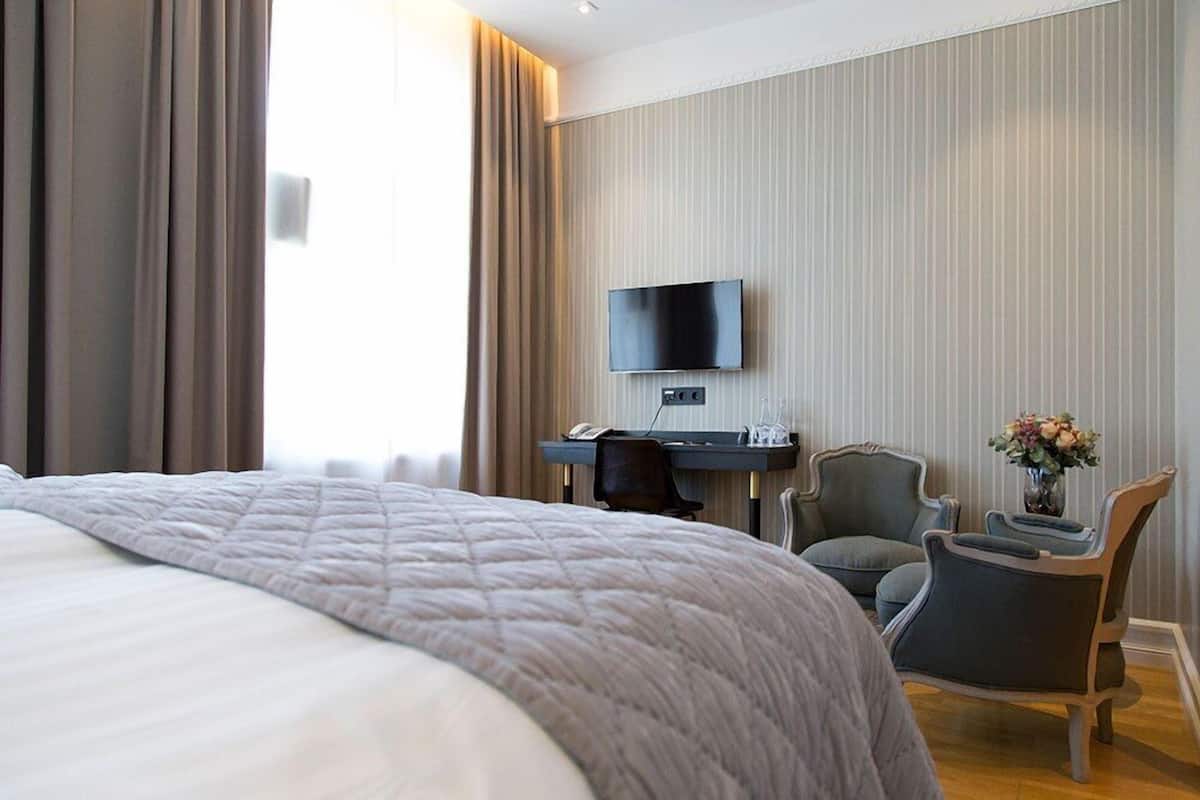 A hotel room with a large bed, a desk with a flat-screen TV, two armchairs, and a vase with flowers.