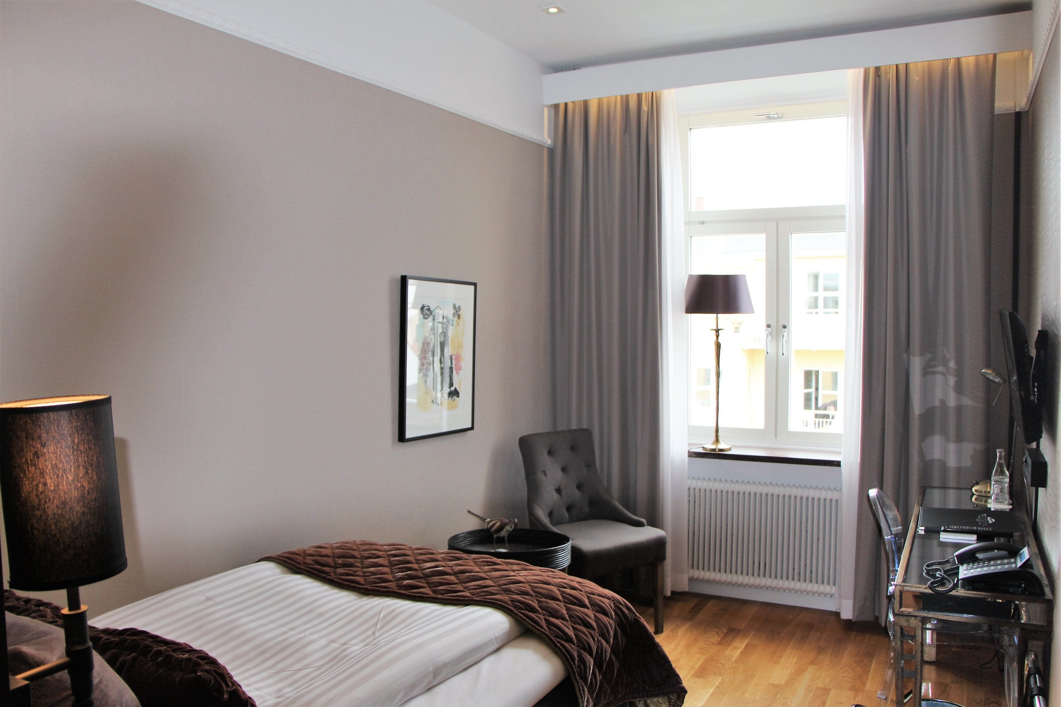 A hotel room with a bed, a chair, a desk with a lamp, a picture on the wall, and a window with curtains.