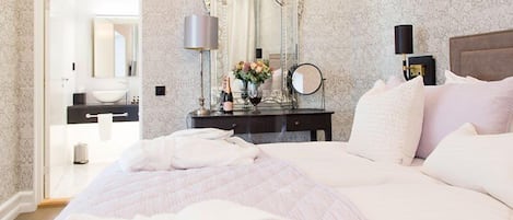 A hotel room with a large bed, a vanity with a mirror, a nightstand with a lamp, and a view of the bathroom.