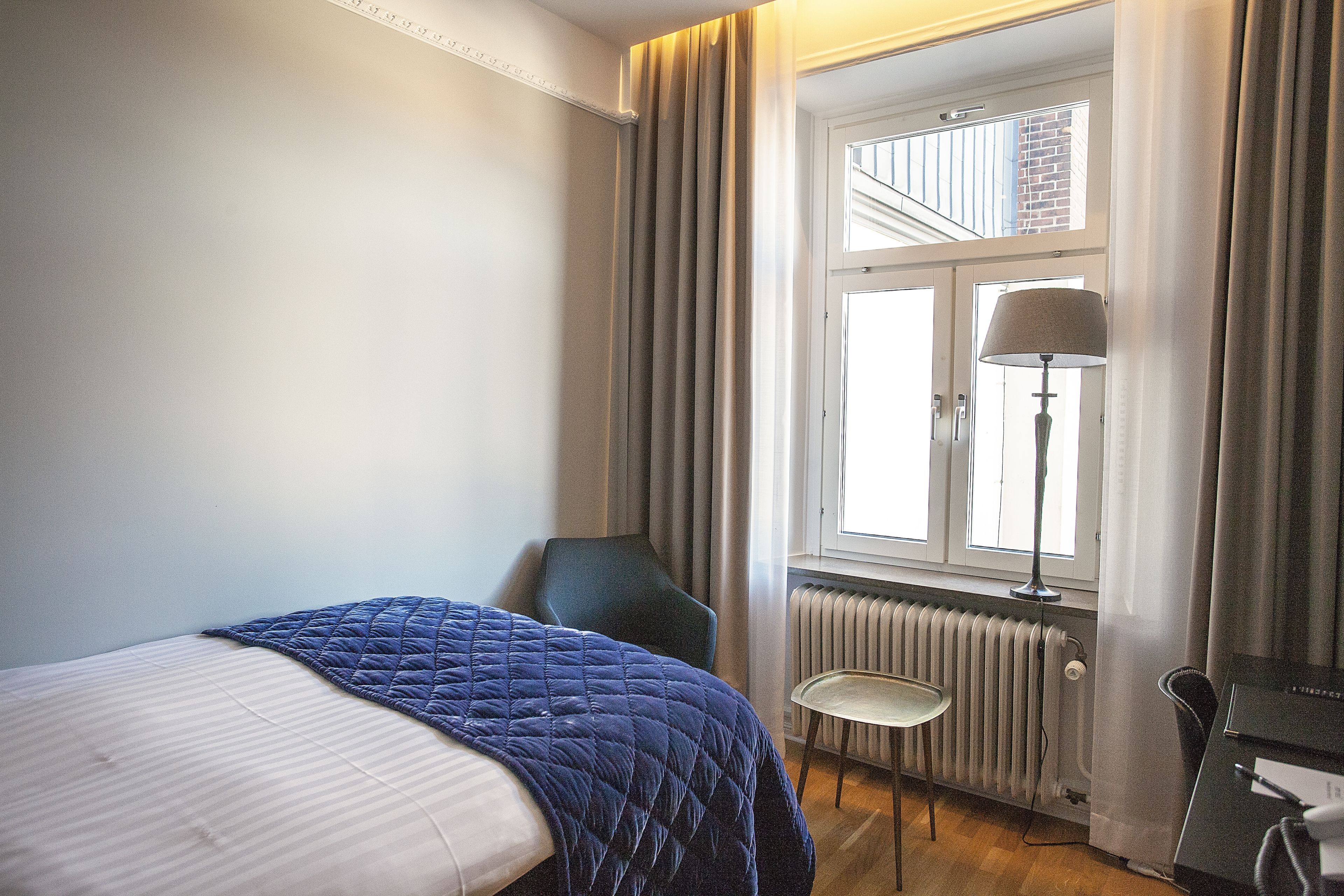A hotel room with a bed, a chair, a desk, a lamp, and a window with curtains.