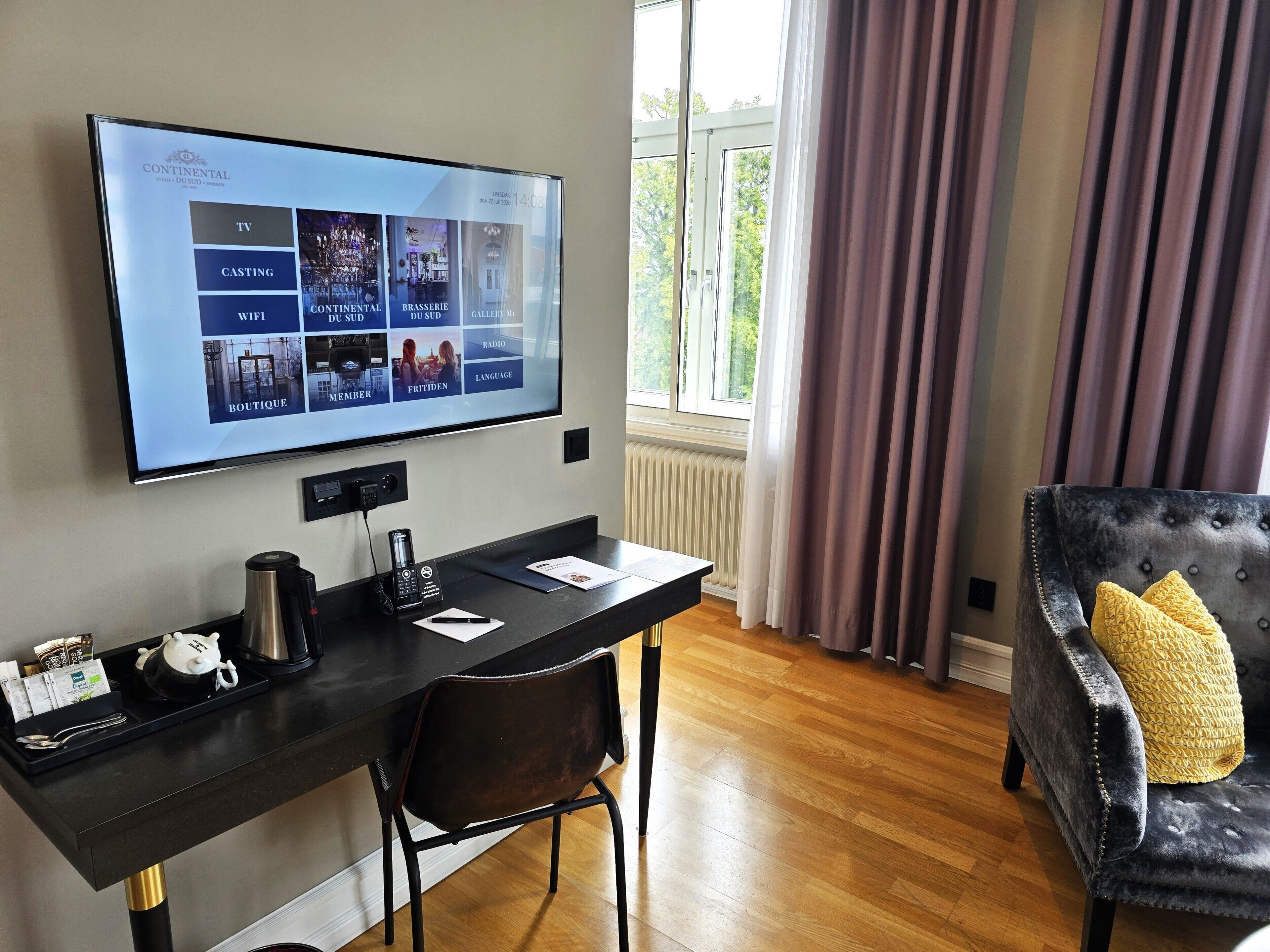 Habitación de hotel con televisión de pantalla plana montada en la pared, un escritorio con silla, un sofá de cuero negro con un cojín amarillo y una ventana con cortinas.