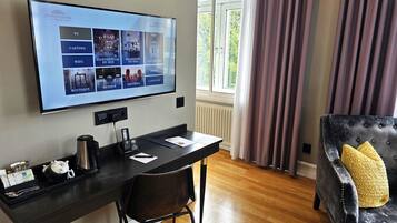 Habitación de hotel con televisión de pantalla plana montada en la pared, un escritorio con silla, un sofá de cuero negro con un cojín amarillo y una ventana con cortinas.