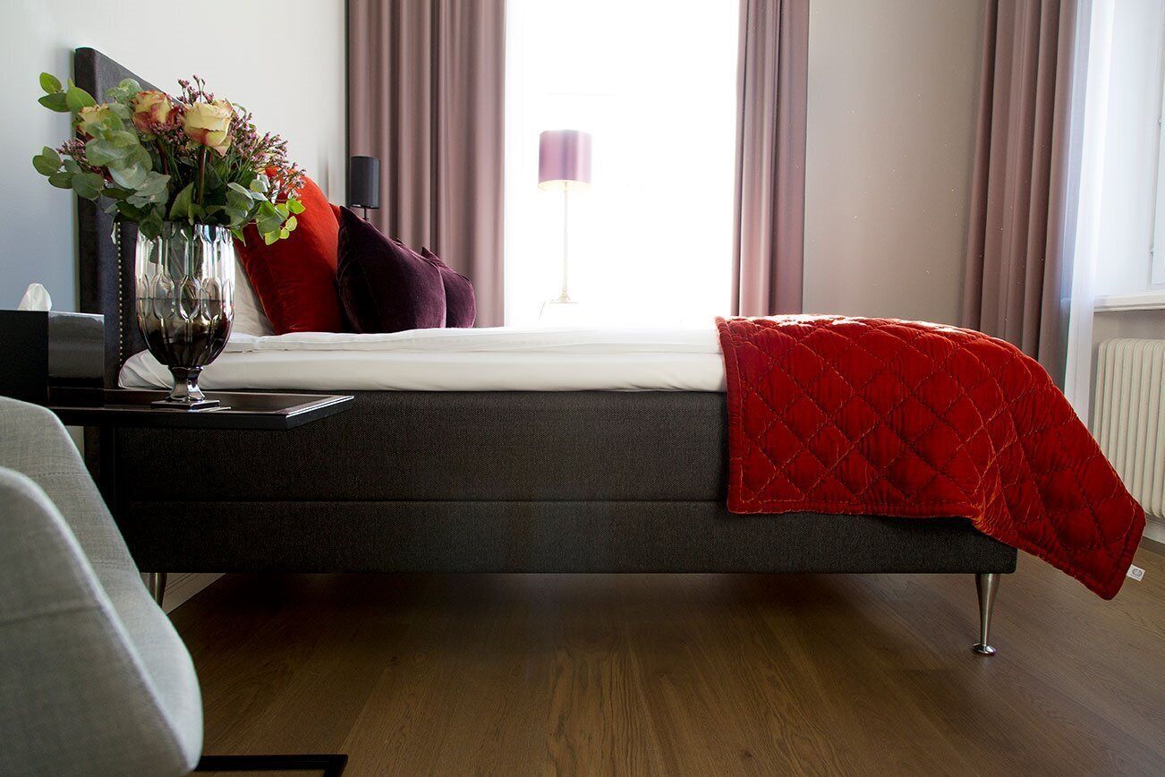 Un dormitorio moderno con una cama color gris oscuro, una colcha acolchada roja y un jarrón de flores en la mesita de noche.
