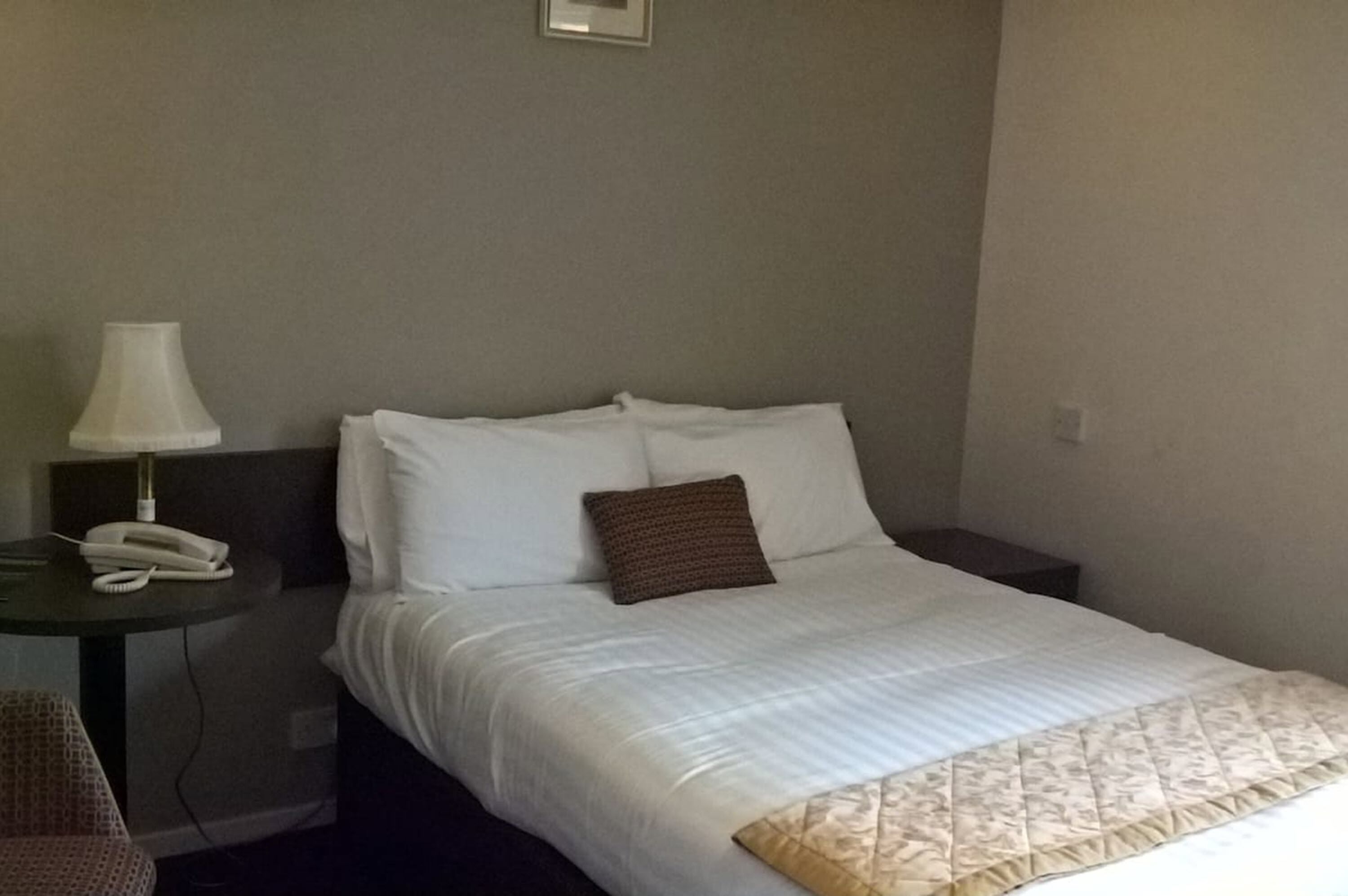 A hotel room with a bed, a bedside table with a lamp, a phone, and a chair.