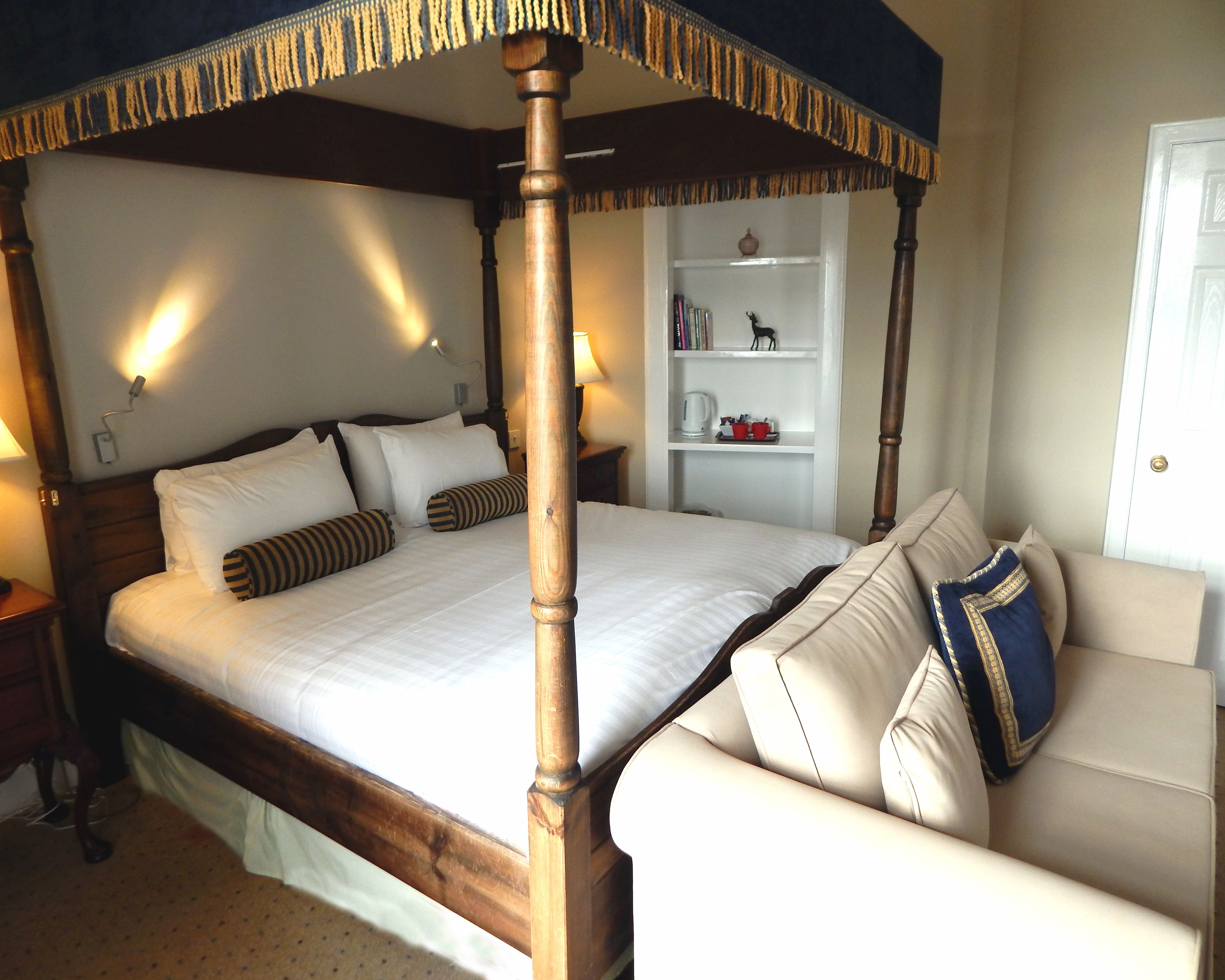 Una cama con dosel de madera, un sofá blanco con cojines estampados, una mesita de noche con lámpara, y una estantería con libros y objetos decorativos.