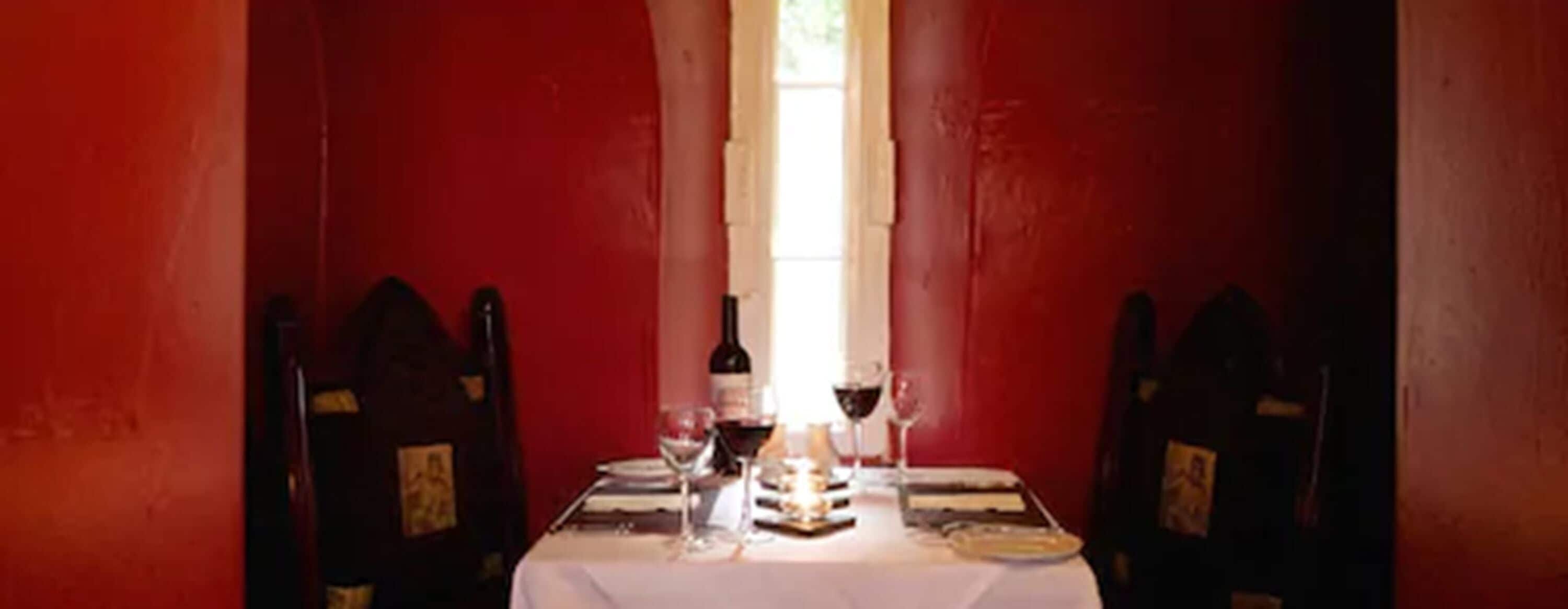 A dining table set with wine glasses, a bottle, and plates, flanked by red walls and wooden chairs.