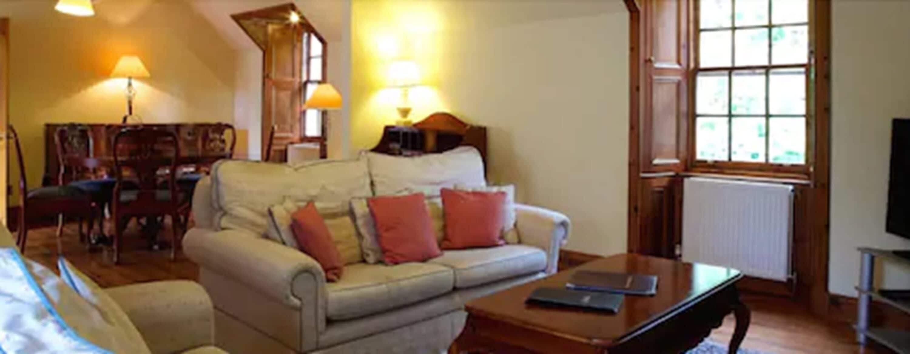 A cozy living room with a sofa, a coffee table, and a television. There is a window with blinds, a lamp, and a chair.