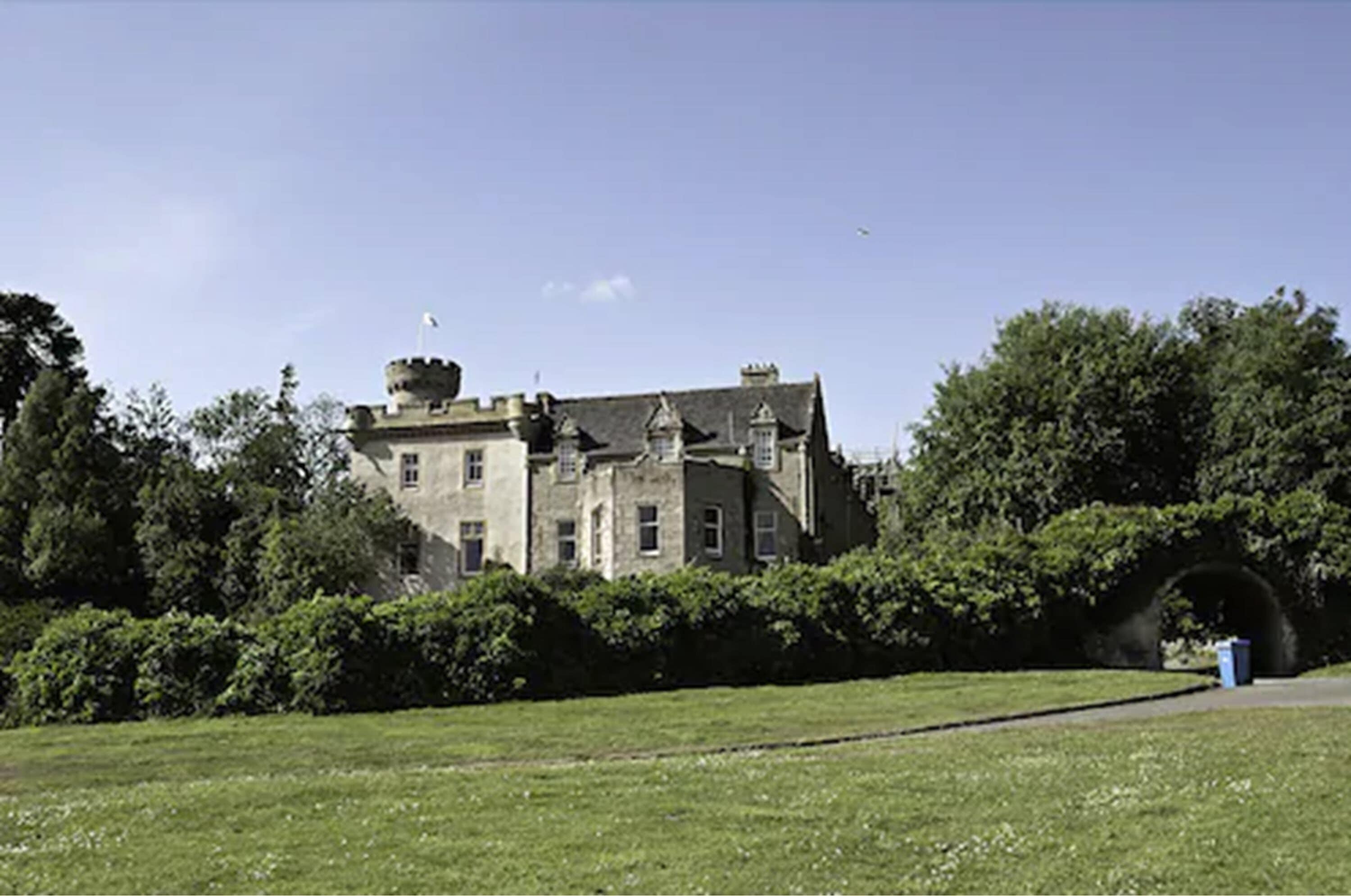 A large, historic building with a prominent tower and multiple chimneys, surrounded by lush greenery and a clear sky.
